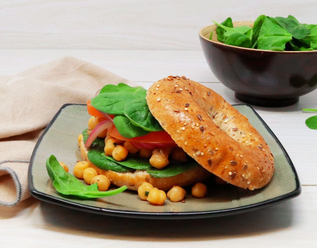 Close up shot of Country Harvest 14 Grain Bagel topped with chickpeas, spinach leaves, red onion and slices of red peppers