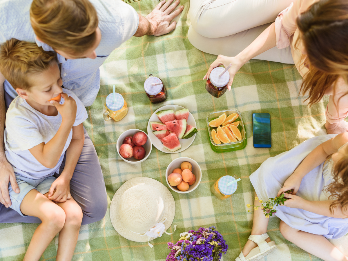 Family of 4 sitting on a green picnic blanket with fruits in the center